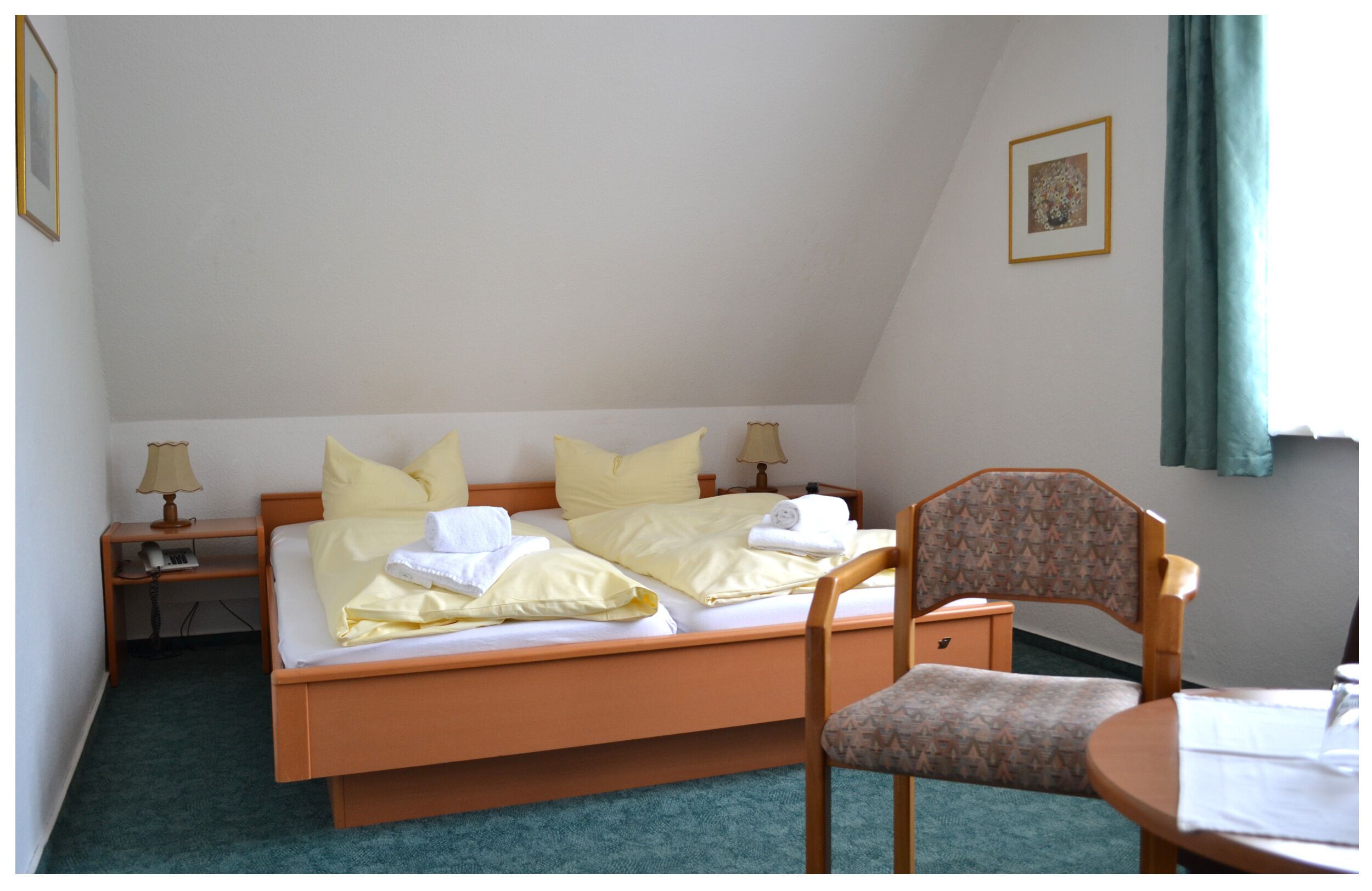 A single bed with a wooden frame, a bedside table with a lamp, a chair, and a small table with a tablecloth.