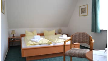 A single bed with a wooden frame, a bedside table with a lamp, a chair, and a small table with a tablecloth.