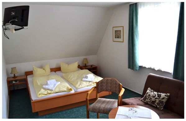 A hotel room with a bed, two bedside tables, a chair, a small table, a TV mounted on the wall, and a window with curtains. - Zum Harzer Jodlermeister (Thale)