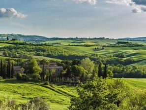A rural landscape with rolling hills, a house surrounded by trees, and a distant village.