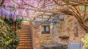 A rustic outdoor patio with a stone wall, wooden steps, a table with chairs, and a hanging light fixture. - Bertusi Source- The Sunset (Pienza)