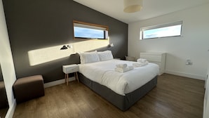 A modern bedroom with a large bed, bedside tables, a nightstand, a small stool, and two windows.