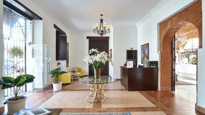 A hotel lobby with a reception desk, a chandelier, a glass coffee table with a vase of flowers, a yellow armchair, and a large window with a view of the street. - Relaxing Getaway in a Colonial Hotel
 (Zona colonial)