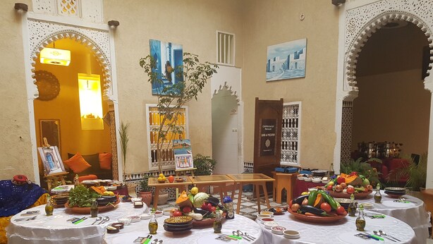 Un comedor con varias mesas redondas cubiertas con manteles blancos, decorado con frutas y verduras frescas, y rodeado de arcos blancos ornamentados.