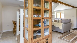 A dining area with a wooden cabinet containing dishes and glasses, a dining table with chairs, and a grey sofa in the background. - Woodcut Cottage, Free WiFi, Rural Location (Evesham)