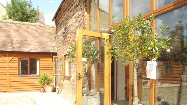 A wooden building with a glass door entrance, a small tree in a planter, and a window with a view of the surrounding area. - Woodcut Cottage, Free WiFi, Rural Location (Evesham)