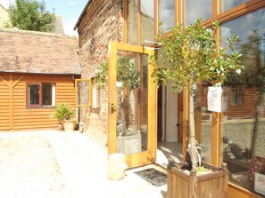 A wooden building with a glass door entrance, a small tree in a planter, and a window with a view of the surrounding area. - Woodcut Cottage, Free WiFi, Rural Location (Evesham)