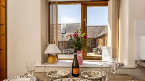 Ein Esstisch ist gedeckt mit Tellern, Gläsern und einer Weinflasche, mit Blick aus dem Fenster auf ein Gebäude und einer Blumenvase.