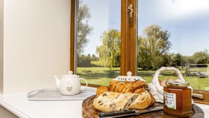 En frukostuppsättning med bröd, croissanter, en tekanna och honung, uppdukad på en träbricka vid ett fönster med utsikt över träd och en byggnad.