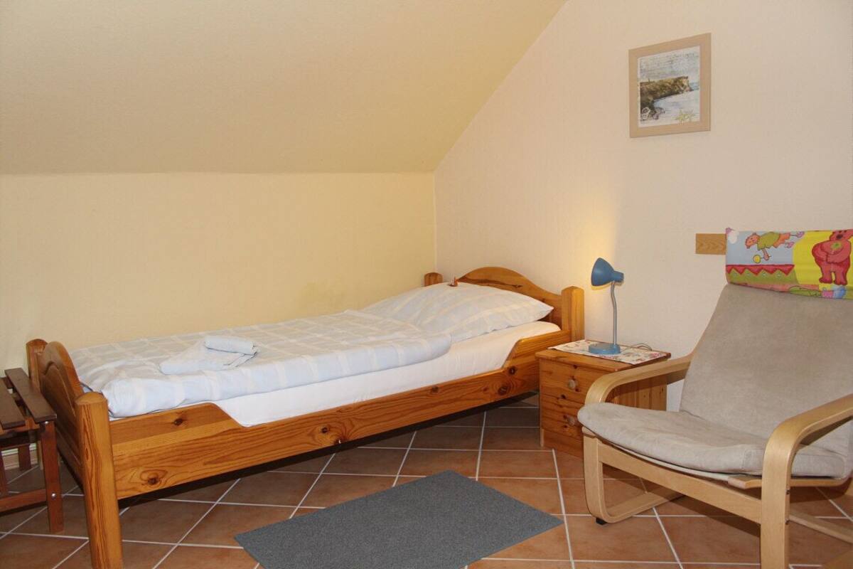 Una cama individual con estructura de madera, una mesita de noche con lámpara, una silla y una alfombra pequeña sobre piso de baldosas.