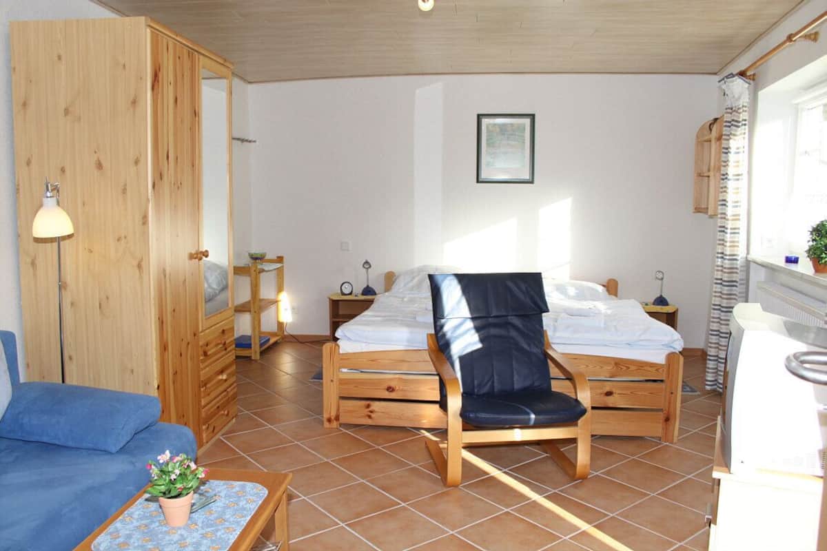 Una habitación con una cama, una silla, un sofá, una mesita con una planta y una ventana con cortinas.