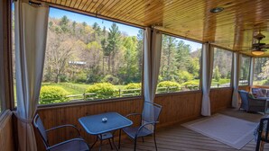 A covered outdoor area with a table and chairs, large windows with curtains, and a view of a forested landscape. - Firefly Fields | Minutes to Blue Ridge Parkway | Hot Tub & Screened Porch (Fleetwood)