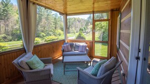 A covered outdoor area with wicker furniture, a table, and a view of a forested landscape. - Firefly Fields | Minutes to Blue Ridge Parkway | Hot Tub & Screened Porch (Fleetwood)