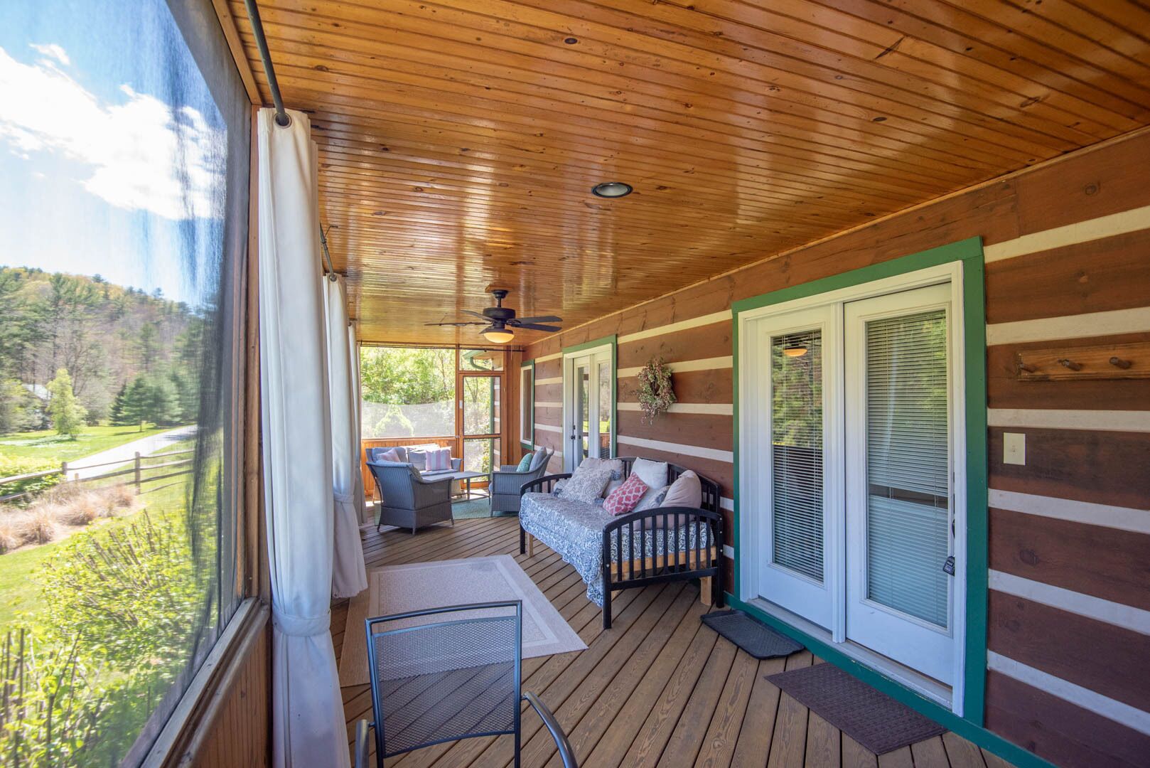 A covered porch with a wooden ceiling, a bed, two chairs, and a view of a green landscape.