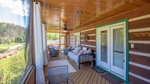 A covered porch with a wooden ceiling, a bed, two chairs, and a view of a green landscape. - Firefly Fields | Minutes to Blue Ridge Parkway | Hot Tub & Screened Porch (Fleetwood)