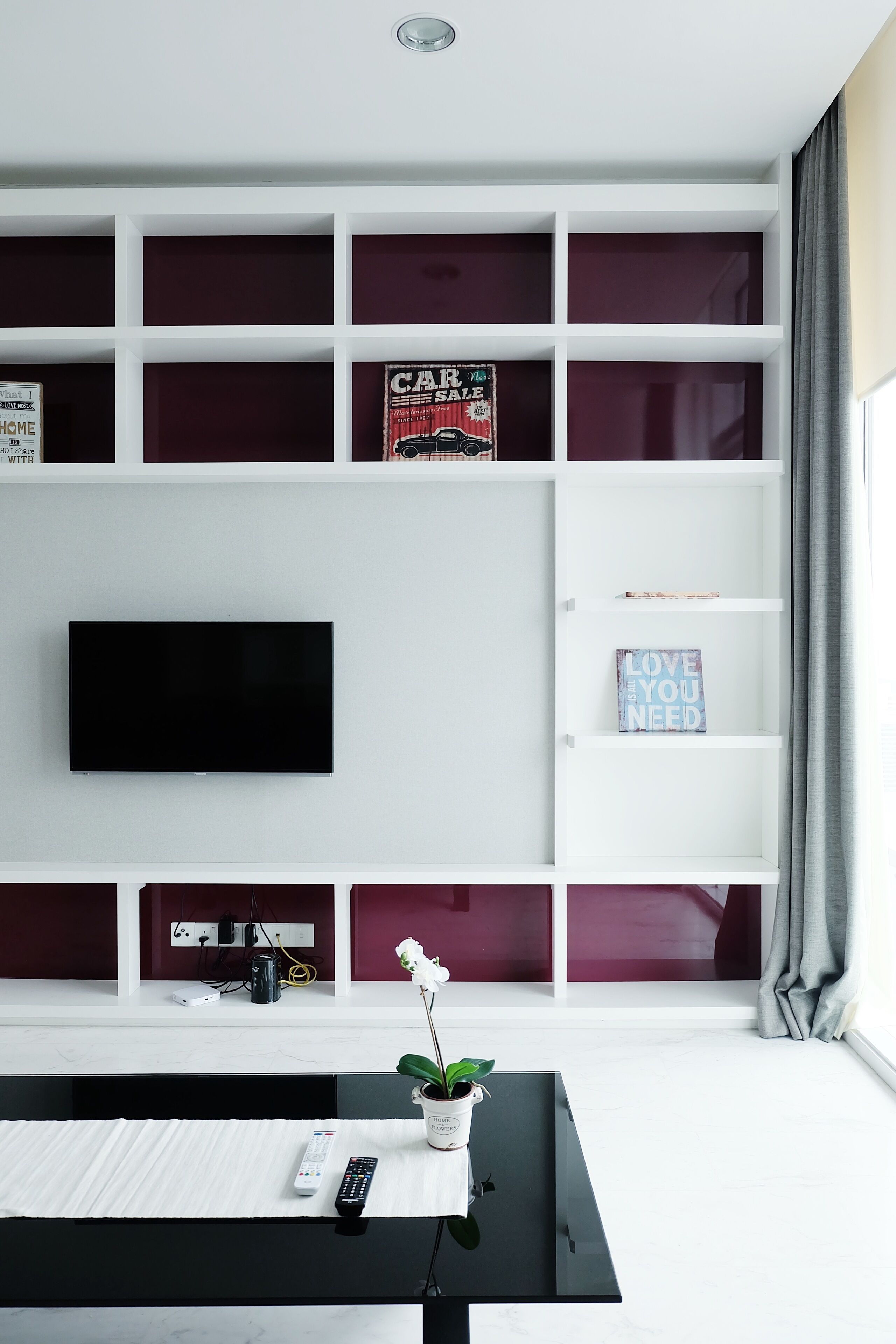 Ein modernes Wohnzimmer mit einem Flachbildfernseher an einer weißen Wand, umgeben von einem weißen Bücherregal mit verschiedenen Gegenständen und einer Topfpflanze auf einem schwarzen Couchtisch.
