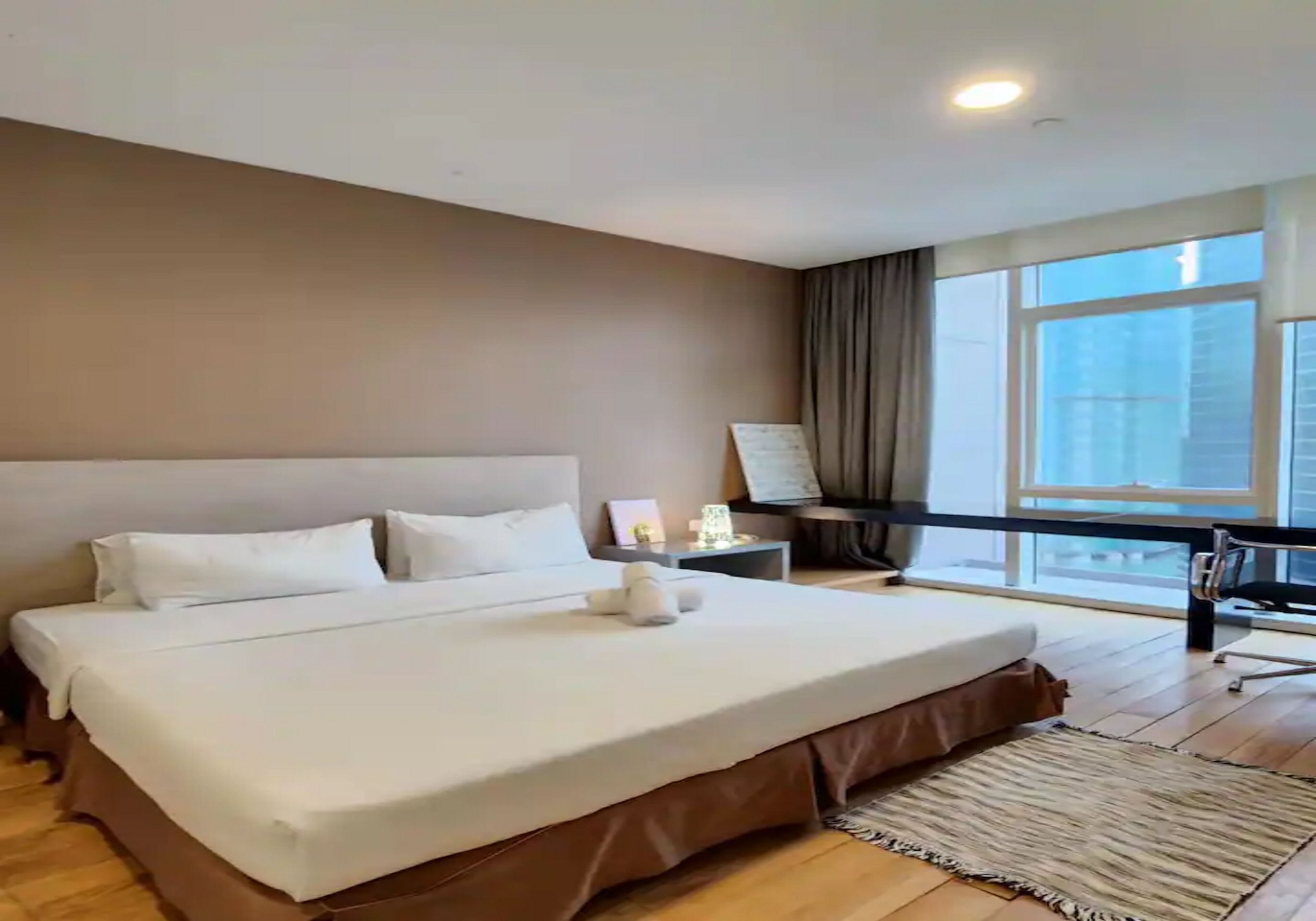 A hotel room with a large bed, a desk, a chair, and a view of the city through large windows.