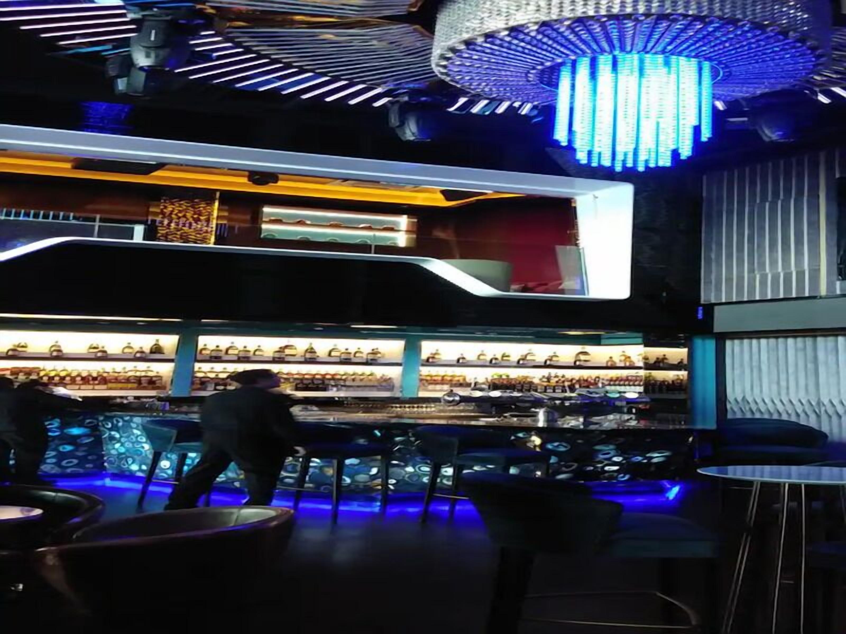 A modern bar area with a high ceiling, blue lighting, and a chandelier.