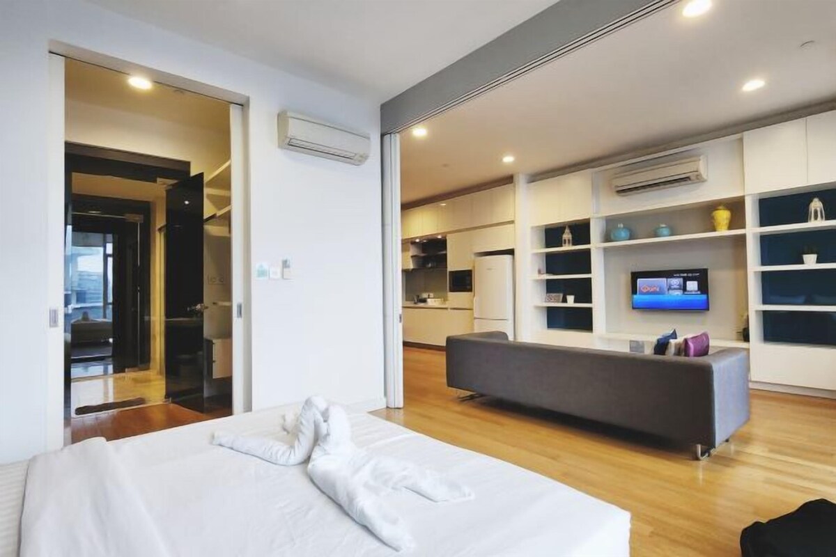 Una habitación de hotel moderna con una cama, un sofá gris, un televisor y estantes empotrados decorados con adornos.