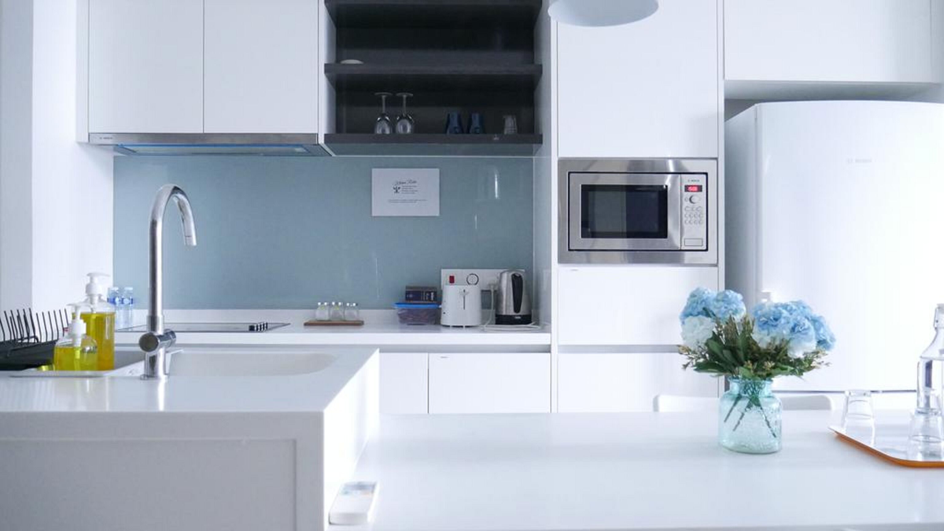 A modern kitchen with white cabinetry, a stainless steel microwave, a kettle, and a vase with flowers on the counter.