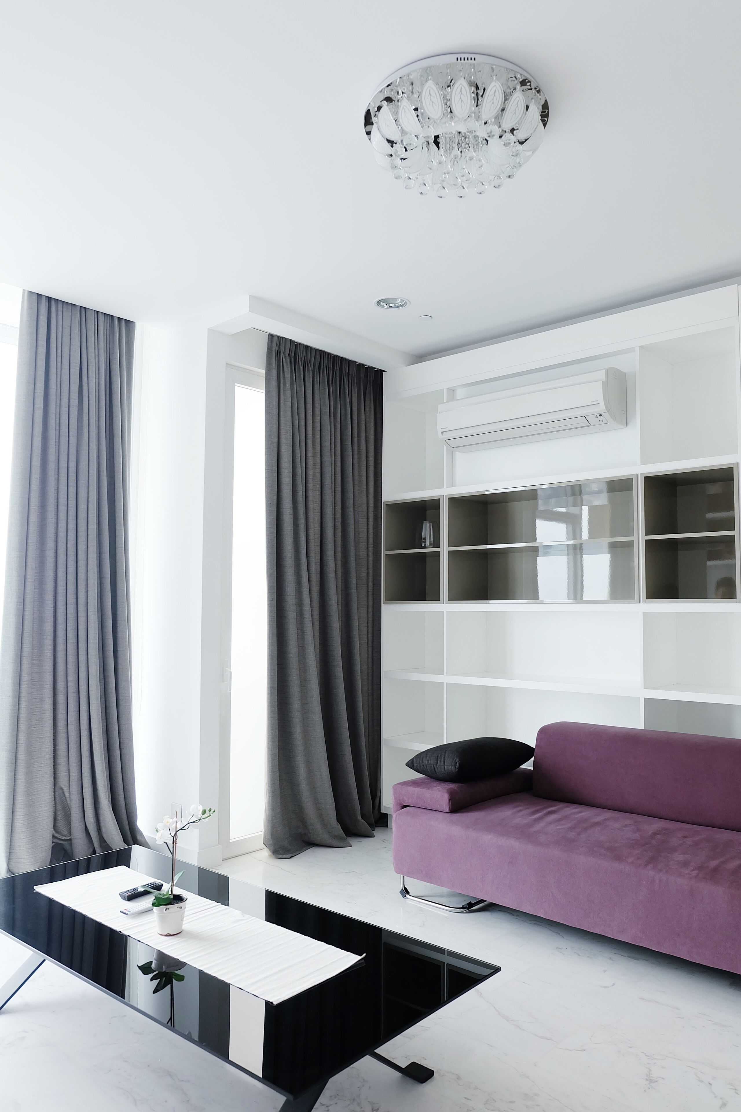 A modern living room with a glass coffee table, a purple sofa, and a white shelving unit.