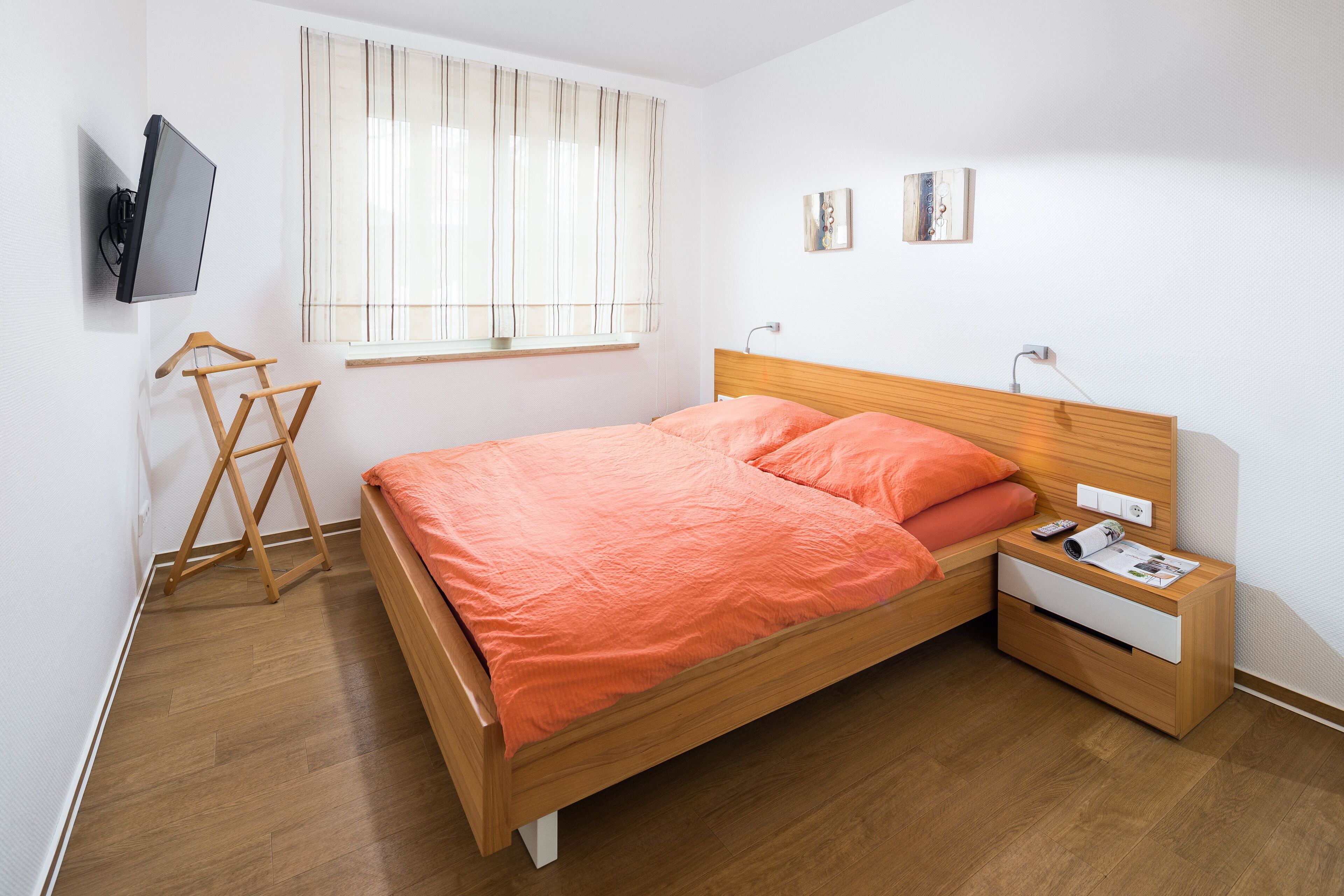 Une chambre à coucher avec un lit en bois, une table de chevet, un téléviseur fixé au mur et une fenêtre avec des rideaux.