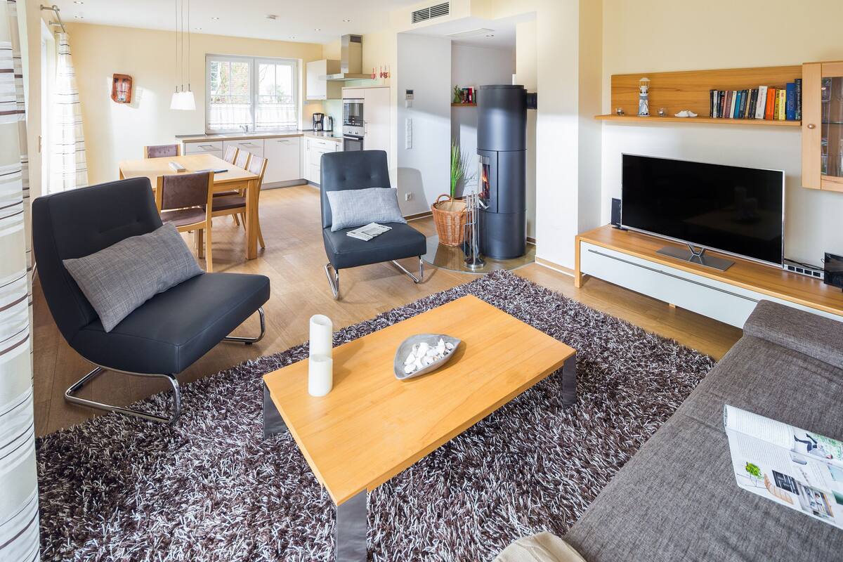 A modern living room with a black sofa, a wooden coffee table, and a TV. There's a kitchen in the background with a stove and cabinets.