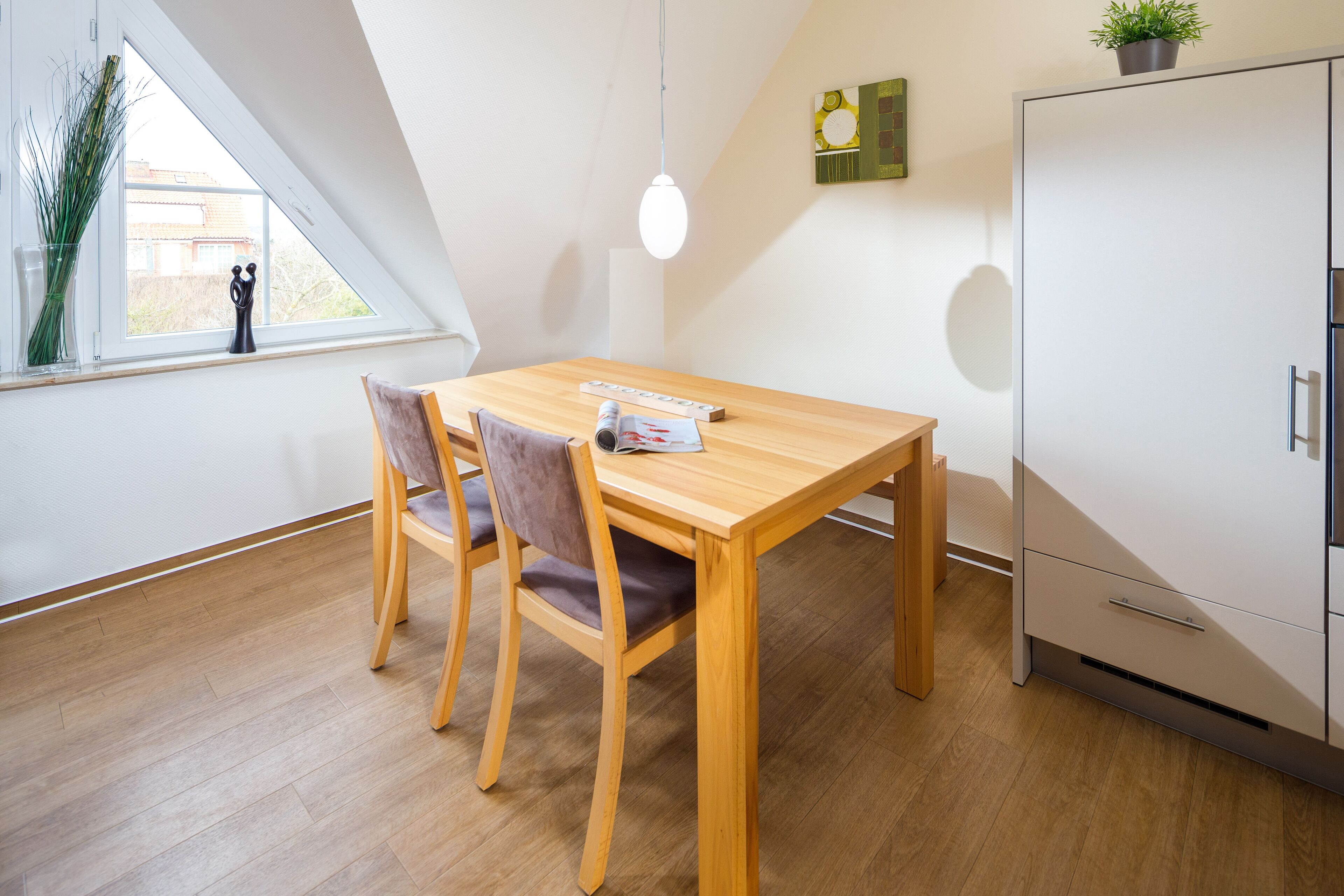 A modern dining area with a wooden table and chairs, a white cabinet, and a window with a view of buildings outside.