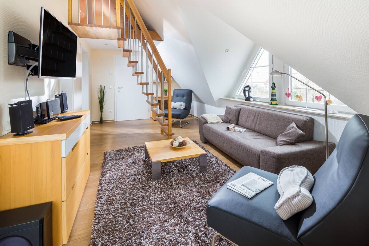A modern loft apartment with a wooden staircase, a grey sofa, a small wooden table, and a TV mounted on the wall.