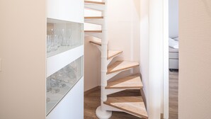A spiral staircase with wooden steps and white walls, leading to a room with a glass cabinet containing wine glasses. - Winkelschiffchen II (Juist)