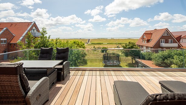 A wooden deck with outdoor furniture, a glass balustrade, and a view of a rural landscape with houses and a hill. - Winkelschiffchen II (Juist)