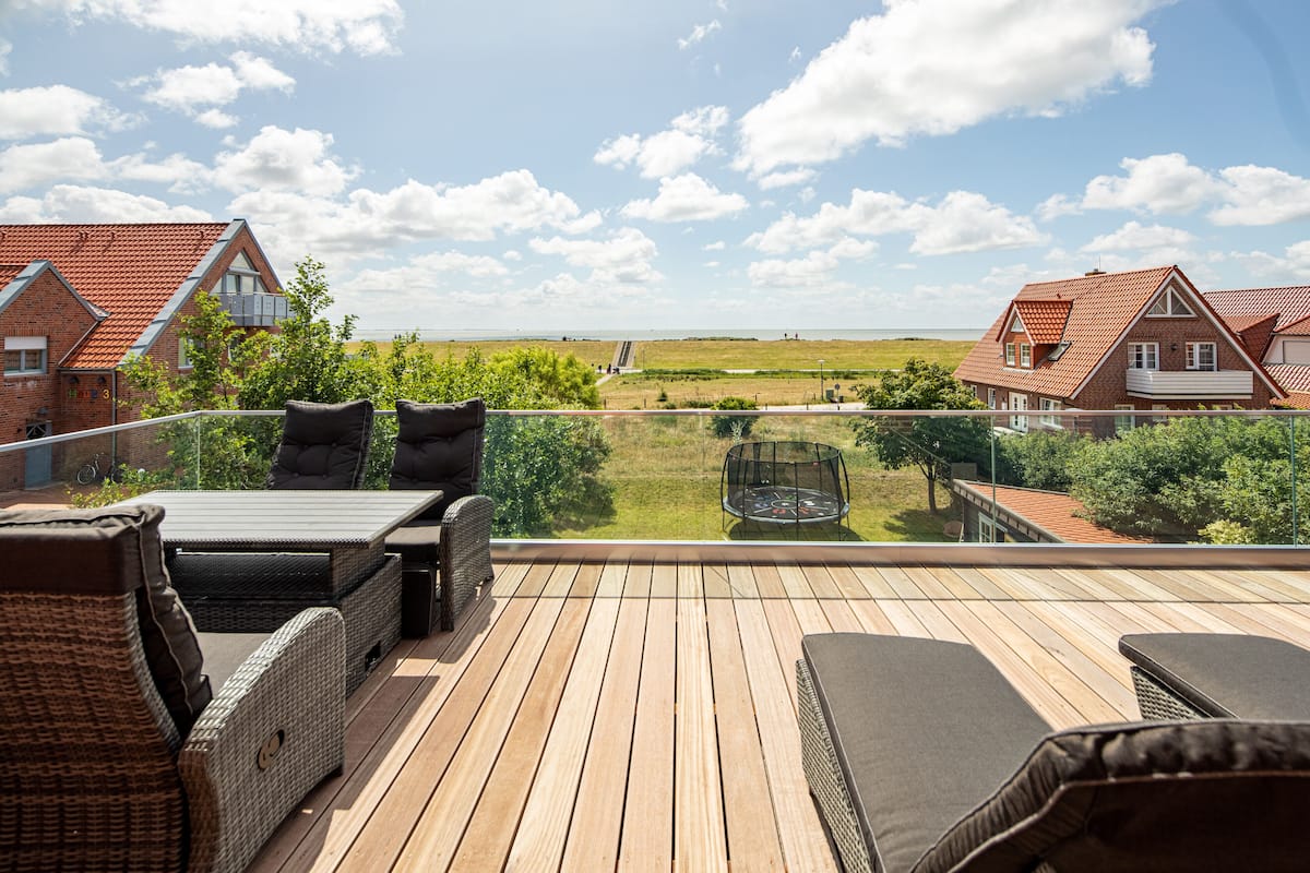 A wooden deck with outdoor furniture, a glass balustrade, and a view of a rural landscape with houses and a hill.