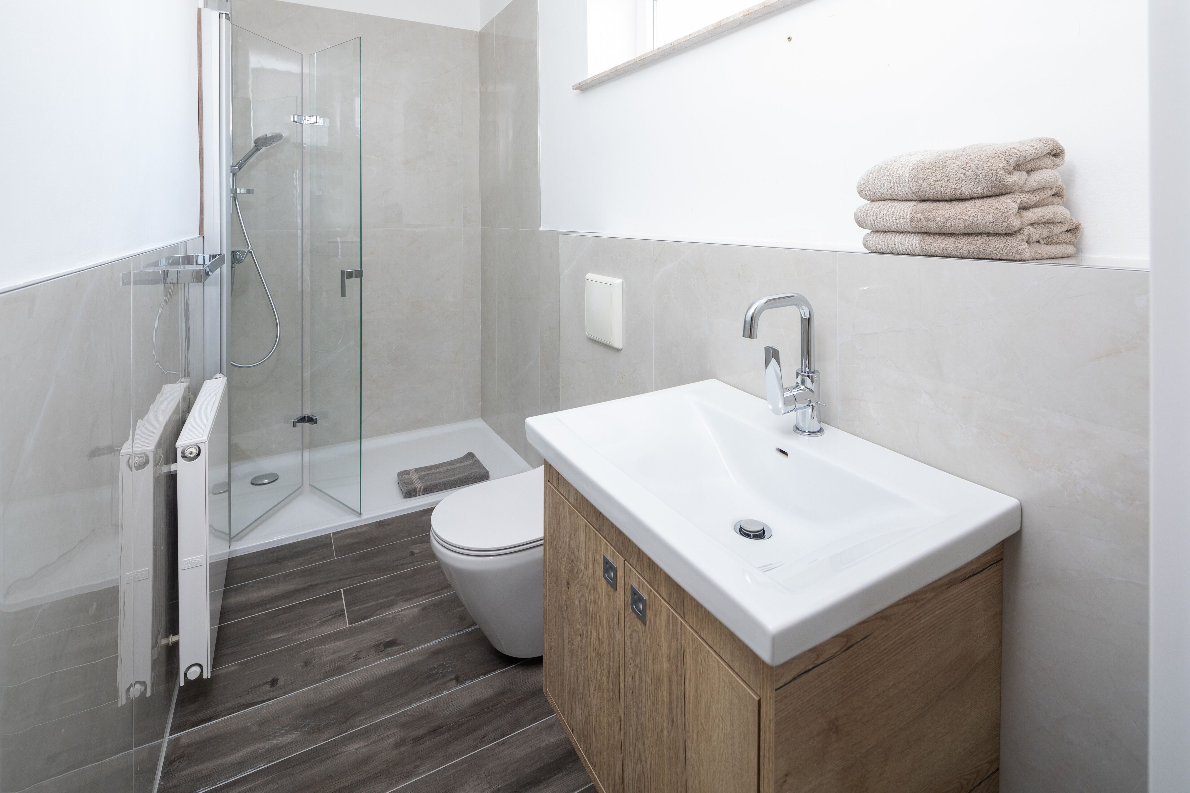 A modern bathroom with a glass shower enclosure, a white toilet, a wooden vanity with a sink, and a stack of towels on the wall.