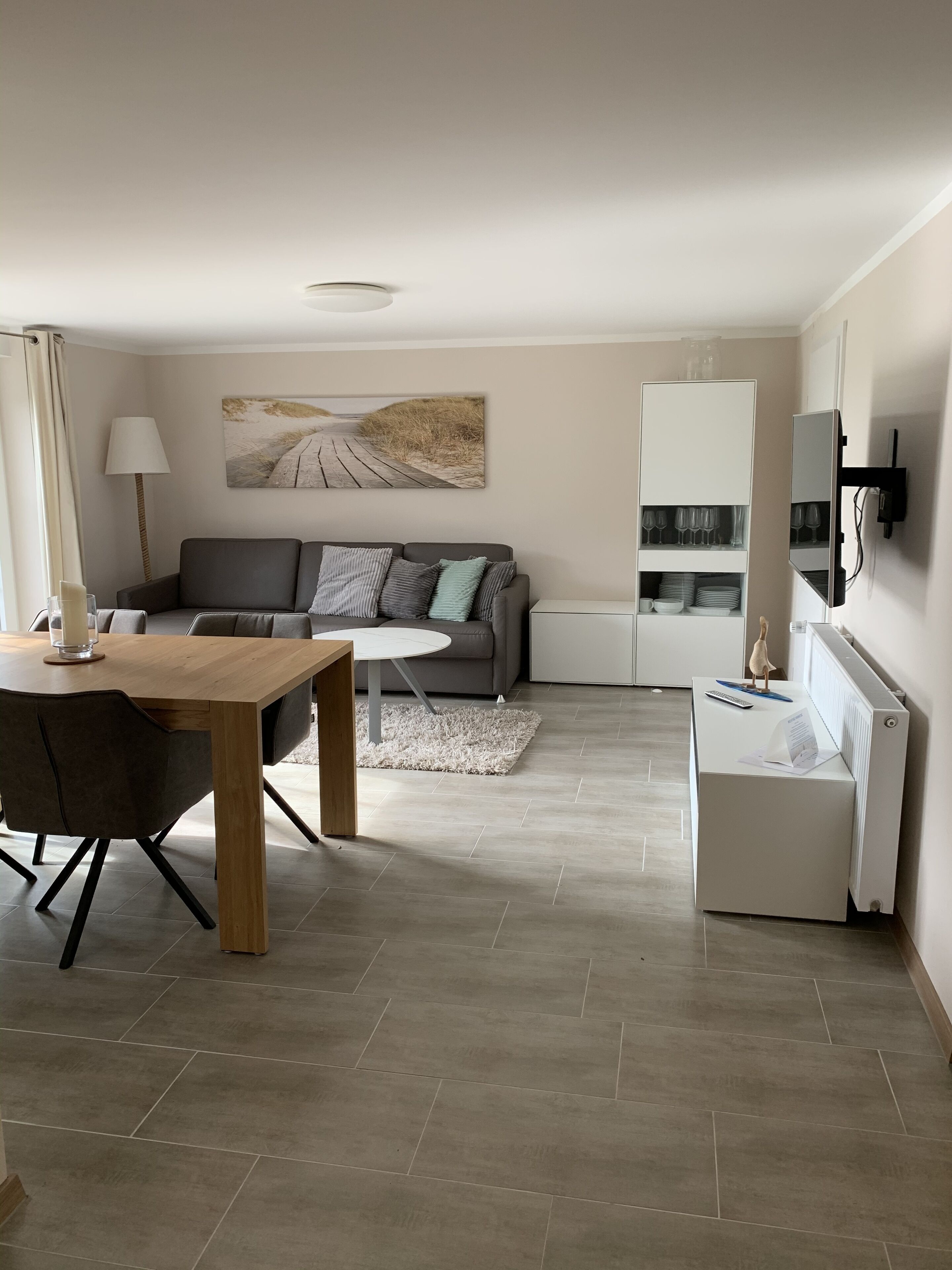Ein modernes Wohnzimmer mit einer Couch, einem Couchtisch und einem Essbereich. An der Wand hängt ein Gemälde, und es gibt eine Lampe sowie einen an der Wand befestigten Fernseher.