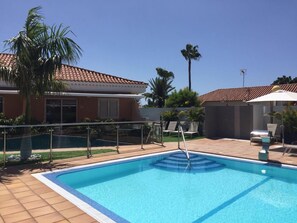 Ein Swimmingpool mit gefliestem Deck, eine Palme und ein Gebäude mit einem Ziegeldach.