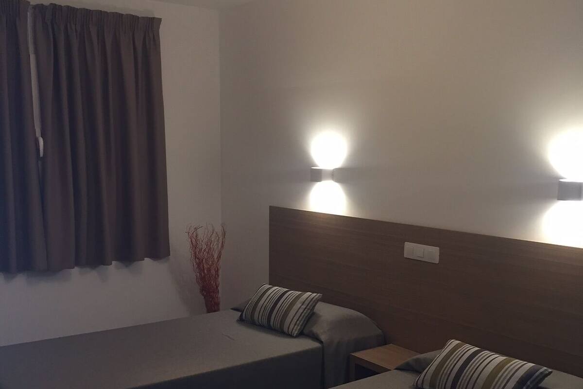 A hotel room with two single beds, a wooden headboard, a nightstand, and a wall-mounted light fixture.
