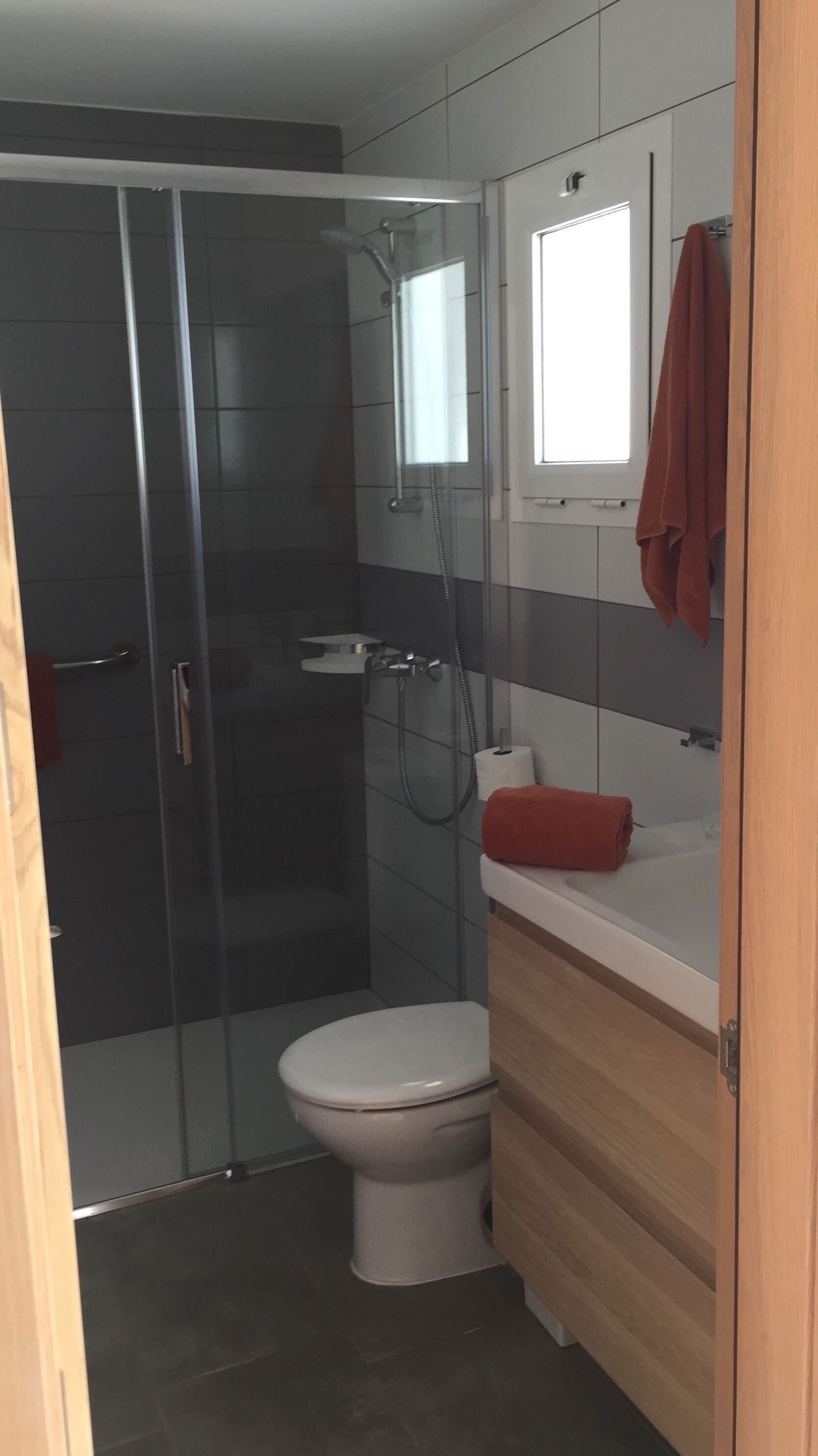 A modern bathroom with a glass-enclosed shower, a white toilet, and a wooden vanity with a red towel.
