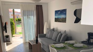 A modern living room with a grey sofa, white chairs, a dining table set for two, and a kitchen area with a stove. - Los Leones Bungalows (San Bartolomé de Tirajana)