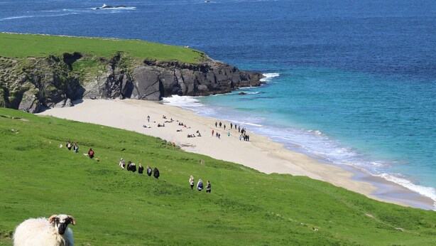 A beach with people walking and sitting, a grassy hill, and a rocky coastline. - Great Blasket Island Accommodation (Great Blasket)