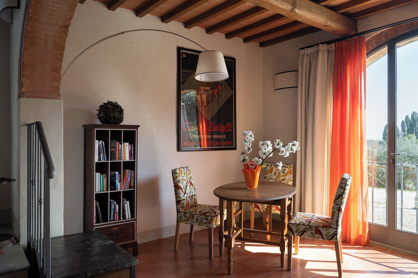 A cozy reading nook with a wooden table, two chairs, a bookshelf, and a large window with curtains.