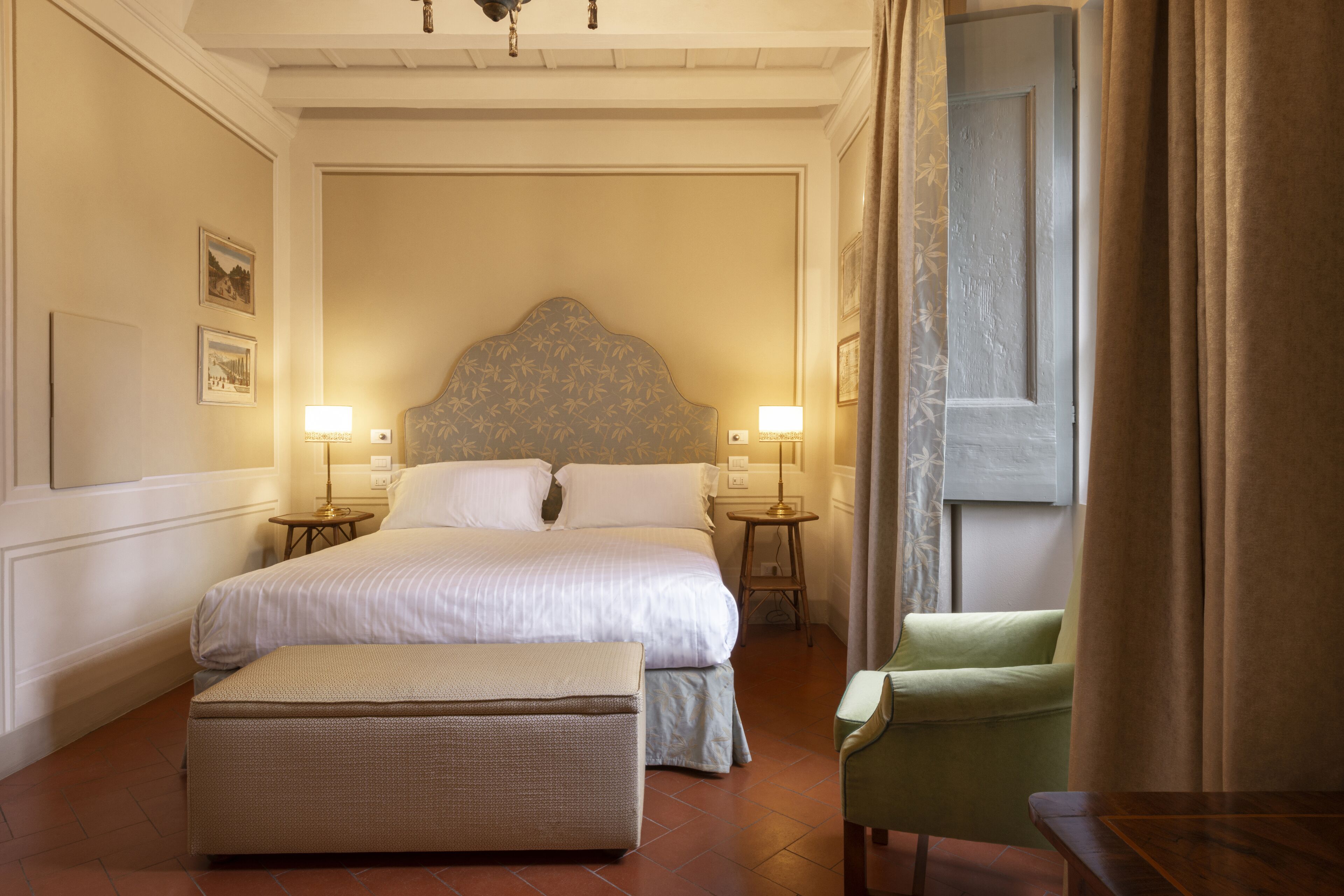 A hotel room with a large bed, two bedside tables, a green armchair, and a small table.