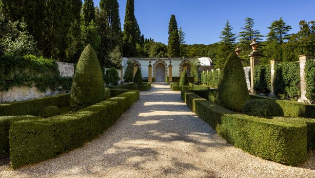 Un jardin à la française, avec des haies taillées avec soin, un chemin de gravier et de grands arbres.