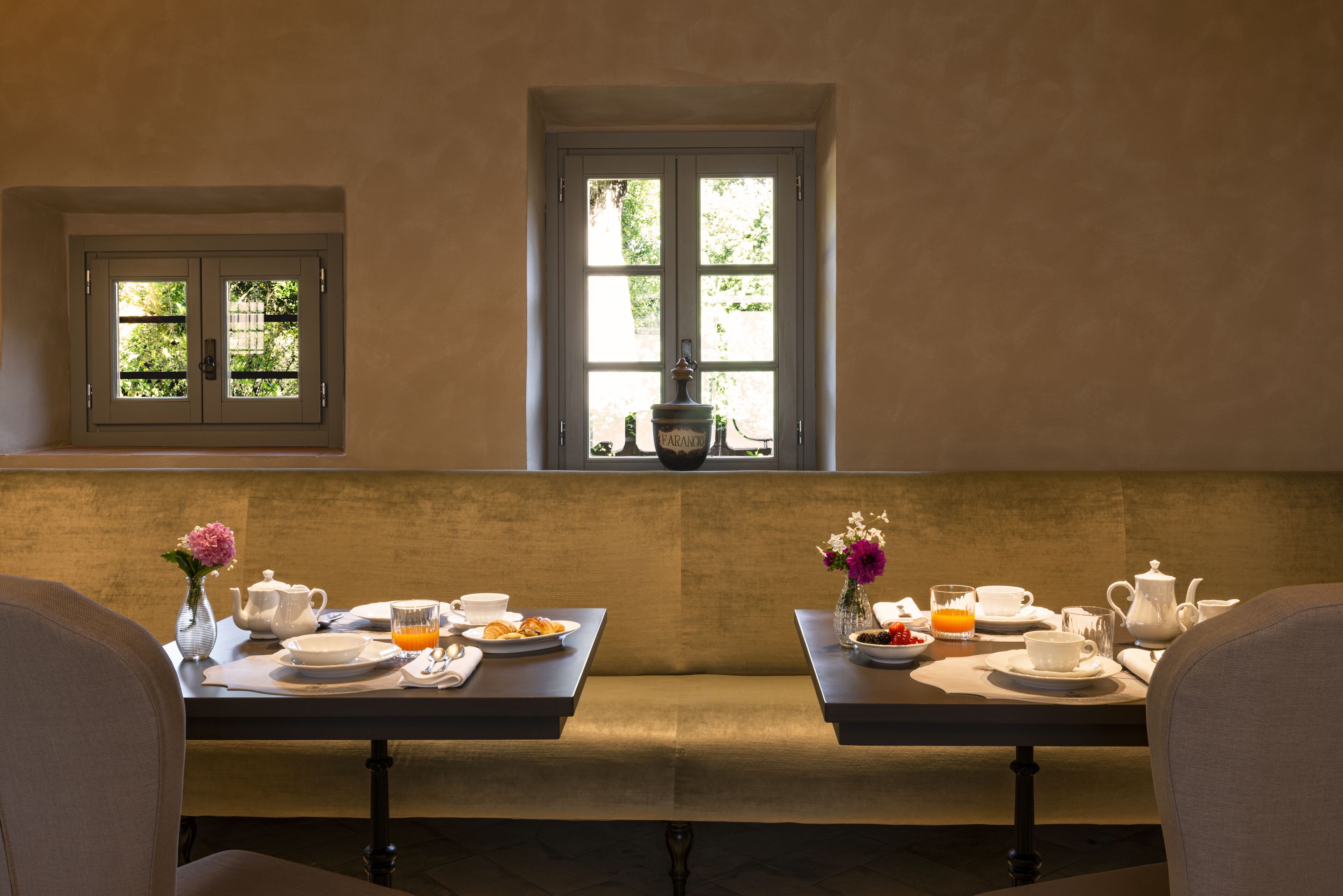 A dining area with two tables set for breakfast, a window with a view of greenery, and a small window with a view of a building.