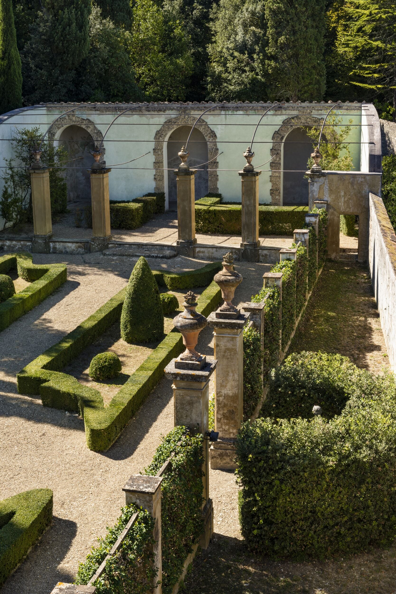 Un jardín formal con setos geométricos, urnas de piedra y estructuras arqueadas.