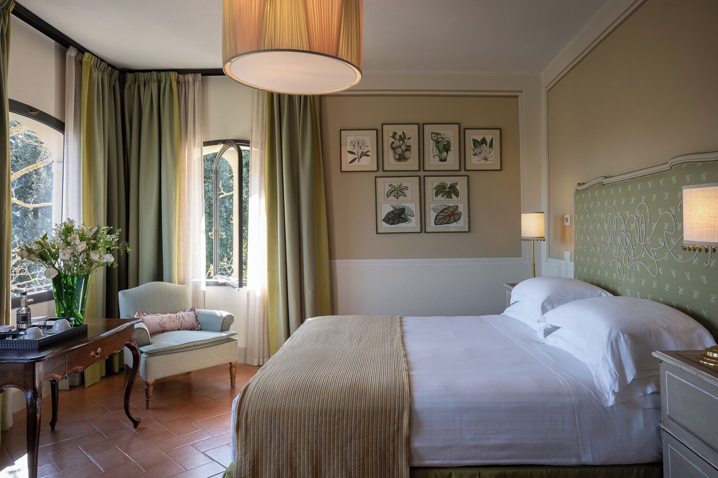 Ein Hotelzimmer mit einem großen Bett, einem Schreibtisch mit Stuhl, einer Blumenvase und gerahmten Pflanzenbildern an der Wand.