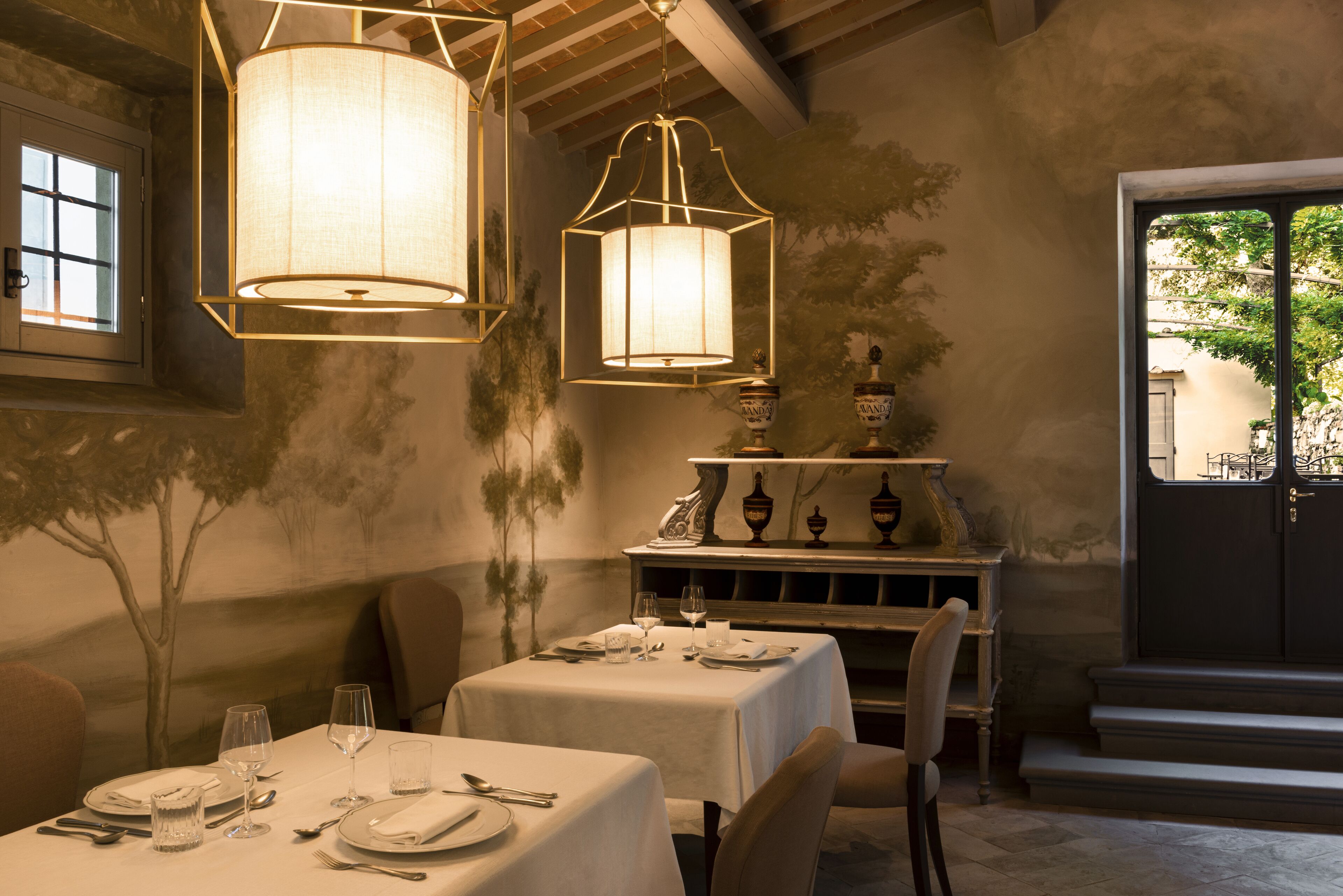 A dining area with tables set for four, each with plates, cutlery, and glasses. There are two hanging lamps above the tables.