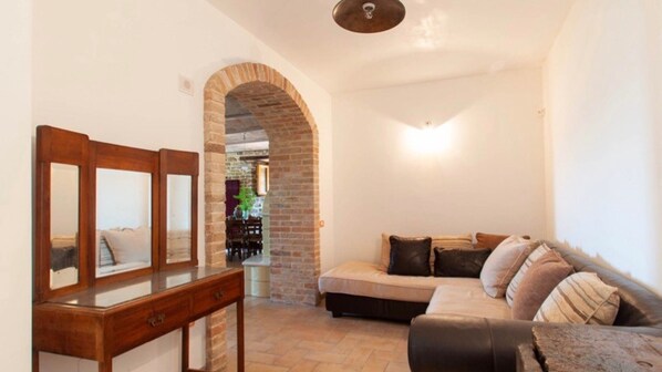 A living room with a sofa, a wooden side table, and a mirror. There is a brick wall and a doorway leading to another room.