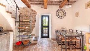 A rustic interior with a stone wall, a wooden ceiling, a dining table with chairs, a metal rack with baskets, and a clock on the wall.