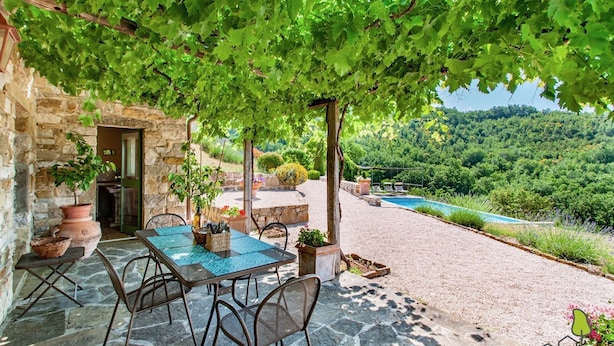 A stone building with a covered patio, outdoor seating, and a view of a pool and forested hills.