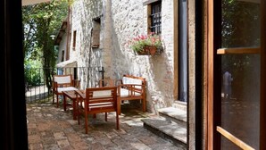 Un bâtiment en pierre avec une table et des chaises en bois à l’extérieur, une fenêtre ornée de fleurs et un portail en métal.
